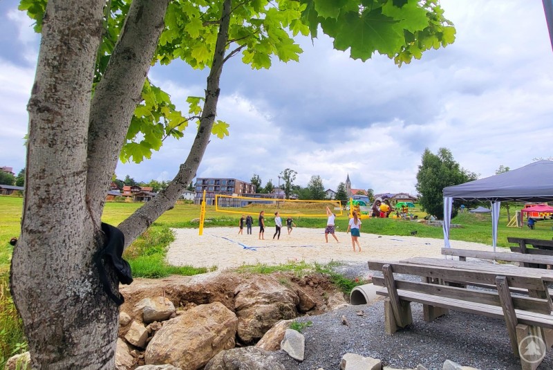 Die wunderschöne Szenerie am Naturbadeweiher sowie eine super entspannte Atmosphäre in der BARdeweiher lud den ganzen Tag über dazu ein, die Seele baumeln zu lassen oder im Rahmen des Woid Summa Graffiti-Kunst, Volleyball, Airbrush-Tattoos, Bungee Run King Kong oder Standup-Paddling auszuprobieren. Kinder und Jugendliche spielen Volleyball auf einem Sandfeld am Naturbadeweiher in Neuschönau. Im Hintergrund sind ein Zeltbereich, bunte Aktionsstationen und eine idyllische Landschaft mit Kirche und Wohnhäusern zu sehen.