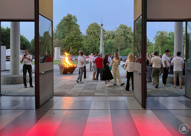 Mit entspannten Gesprächen am Feuerkorb draußen vor der Halle wurde der Sommerabend eingeläutet. Menschen in legerer Kleidung stehen vor geöffneten Hallentüren um einen Feuerkorb, während der Himmel langsam dunkler wird.