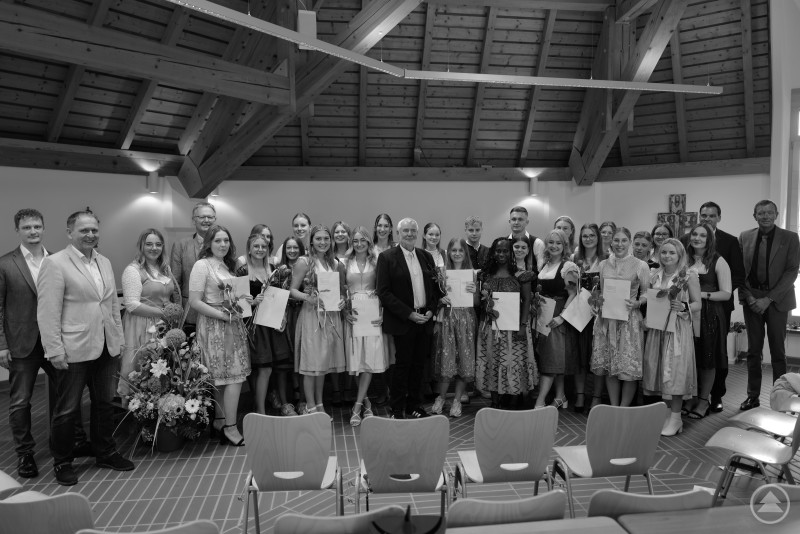 Die 26 Absolventinnen und Absolventen der Pflegeschule Freyung feierten ihren erfolgreichen Abschluss – neun von ihnen erhielten Staatspreise. Eine große Gruppe von Absolventinnen und Absolventen steht in einem festlich geschmückten Saal, hält Urkunden in den Händen und lächelt in die Kamera.