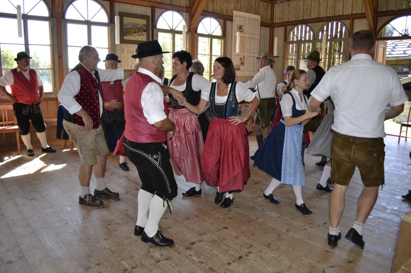 Mehrere Paare in bayerischer Tracht tanzen ausgelassen in einem hellen Holzsaal, begleitet von traditioneller Musik.