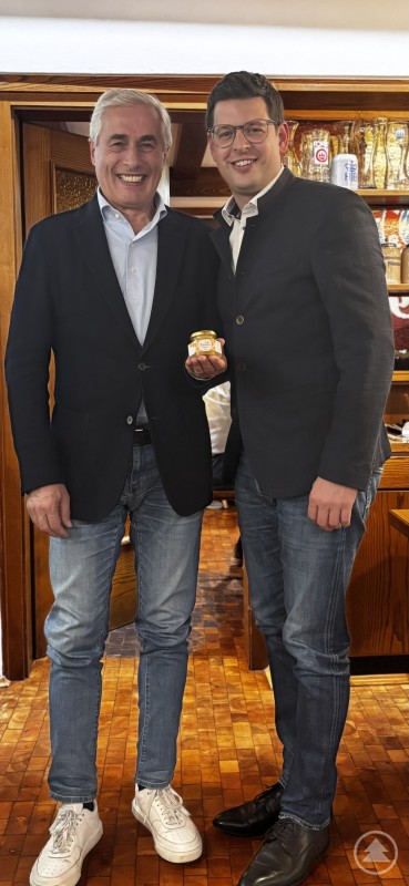 Für Niederdorfs Bürgermeister Günther Wisthaler hatte Bürgermeister Martin Pichler einen süßen Gruß der fleißigen Marktbienen dabei. Zwei Männer stehen nebeneinander in einem holzgetäfelten Raum und lächeln in die Kamera. Einer von ihnen überreicht dem anderen ein kleines Glas Honig als Geschenk.