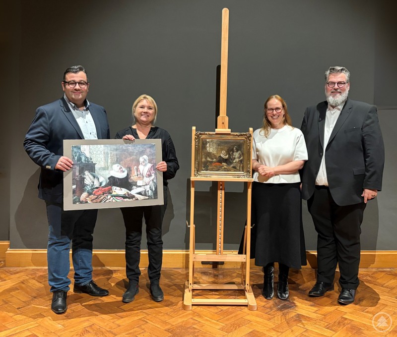 Vier Personen stehen in einem Ausstellungsraum neben einer Staffelei mit einem Gemälde und halten eine Skizze von Ferdinand Wagner in den Händen.