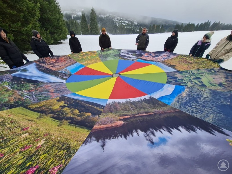 Lernen fand beim deutsch-tschechischen Schulprojekt nicht nur im Klassenzimmer, sondern mitten in der Natur statt – anschaulich, bewegungsreich und nachhaltig. Mehrere Jugendliche und Erwachsene stehen im Kreis auf einer Schneefläche und halten ein großes, buntes Schwungtuch mit Naturmotiven. Im Hintergrund sind verschneite Berge und Wälder zu erkennen.