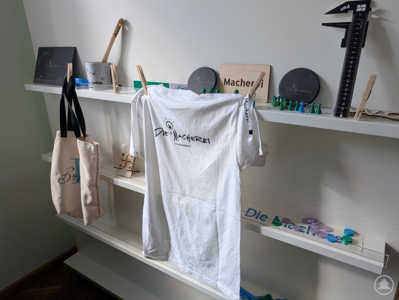 Der Cricut Maker kann mit seinen Aufklebern verschiedene Materialien zieren. Ein weißes Regal mit verschiedenen beschrifteten Materialien wie Tassen, Taschen und T-Shirt sowie Schlüsselanhänger, die mit dem 3D-Drucker erstellt wurden.
