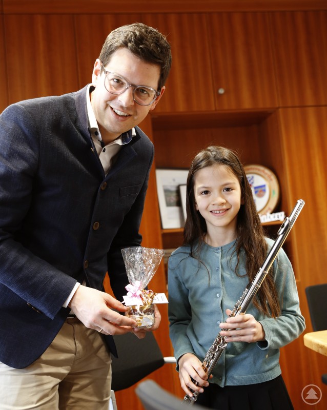 Amelie Hartl schenkte dem stolzen Bürgermeister einen gespielten Blumengruß per Noten – Bürgermeister Martin Pichler revanchierte sich mit Schönberger Honig und Freibadkarten. Ein Mädchen mit Querflöte und ein Mann im Anzug lächeln in die Kamera; sie halten ein Geschenk und stehen in einem Büro mit Holzverkleidung.