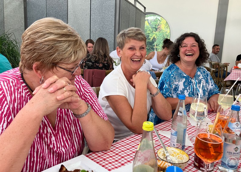 Beste Stimmung beim Sommerfest mit herzhaften Lachen und geselliges Beisammensein. Drei Frauen sitzen an einem Tisch mit rot-weiß karierten Tischdecken, Getränken und Speisen und lachen fröhlich in geselliger Runde.