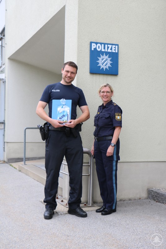PORin Ingrid Grötzinger überreicht das Anerkennungsschreiben des Polizeipräsidiums Niederbayern an Bundespolizist Dominik Saake. Sven Ersek konnte zur Ehrung leider nicht anwesend sein. Ein junger Bundespolizist und eine Polizeibeamtin stehen vor einem Polizeigebäude. Der Beamte hält ein Geschenk und eine Urkunde in der Hand. Beide lächeln in die Kamera.