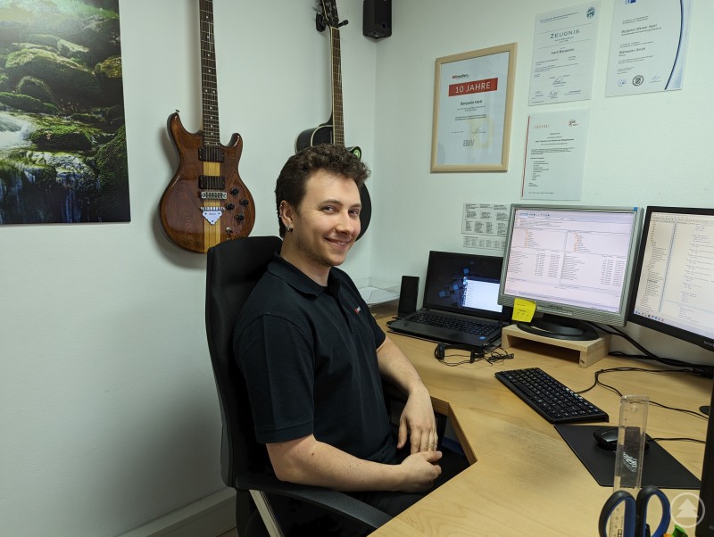 Benjamin Hartl ist das technische Rückgrat von BWmedien: zuverlässig, lösungsorientiert und mit einer guten Portion Humor im Büroalltag. Auf dem Bild ist ein junger Mann an einem Schreibtisch in einem Büro zu sehen. Er sitzt auf einem Bürostuhl, trägt ein schwarzes Poloshirt und lächelt in die Kamera. Vor ihm stehen zwei große Monitore und ein Laptop, alle mit geöffneten Programmier- oder Verwaltungsanwendungen. An der Wand hinter ihm hängen zwei Gitarren, daneben diverse Urkunden und ein Poster mit einem Naturmotiv (Wasserfall im Wald). Die Arbeitsumgebung wirkt technisch und gleichzeitig persönlich gestaltet.