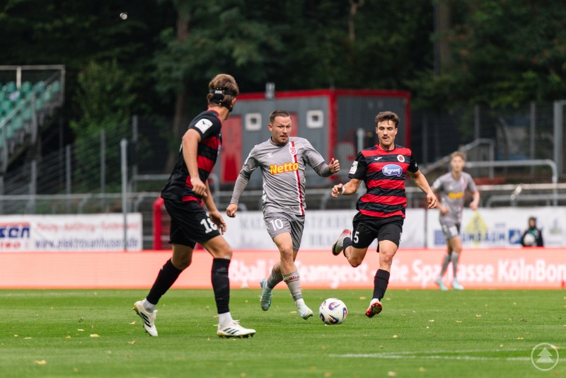 Drei Spieler im Laufduell um den Ball auf dem Spielfeld.