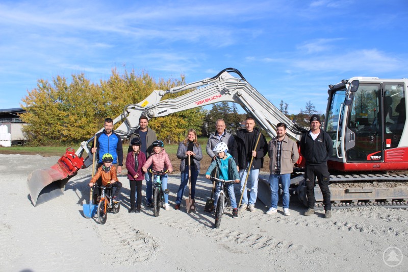 (v.li., hinten) Zsolt Vanczer, Stefan Detter, Markträtin Evi Pöppl, 1. Bürgermeister Robert Grabler, 2. Bürgermeister Johannes Seidl, Bauamtsleiter Maximilian Röckl und der Mitarbeiter der Baufirma mit den bikebegeisterten Kindern (v.li., vorne) Kristof Vanczer, Charlotte Detter, Abigél Vanczer und Jakob Detter beim Spatenstich des Aidenbacher Bikeparks. Eine Gruppe von Erwachsenen und Kindern steht vor einem Bagger auf einer Baustelle. Die Kinder sitzen auf Fahrrädern und tragen Helme, während die Erwachsenen Spaten halten. Im Hintergrund sind herbstlich gefärbte Bäume zu sehen.