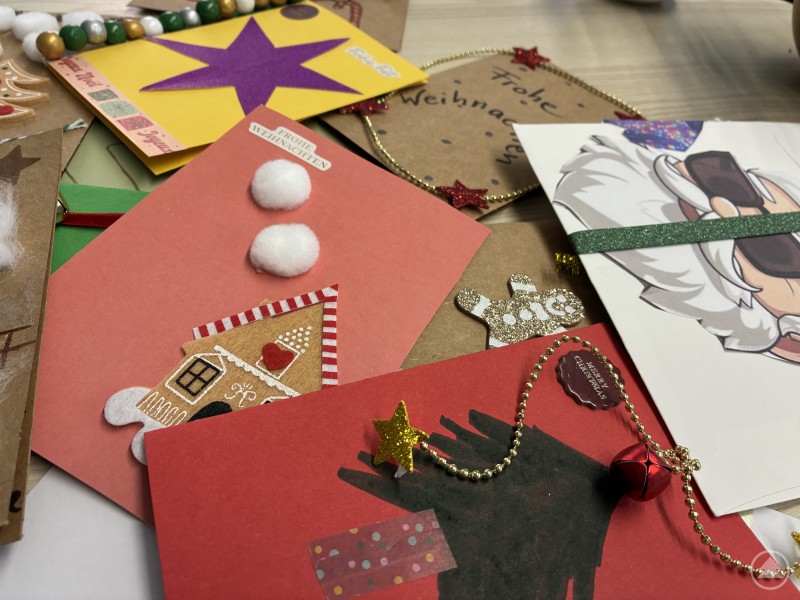 Handgeschriebene Botschaften der Verbundenheit: Briefe, Karten und Zeichnungen der Aktion „Herzenspost – Worte, die verbinden“ warten darauf, Freude zu schenken. Mehrere bunte und liebevoll gestaltete Weihnachtskarten liegen auf einem Tisch. Einige sind mit Glitzer, Sternen, Perlenketten und kleinen Anhängern verziert. Die Karten tragen handgeschriebene Grüße und Motive wie Weihnachtsbäume, Häuser und Schneeflocken.