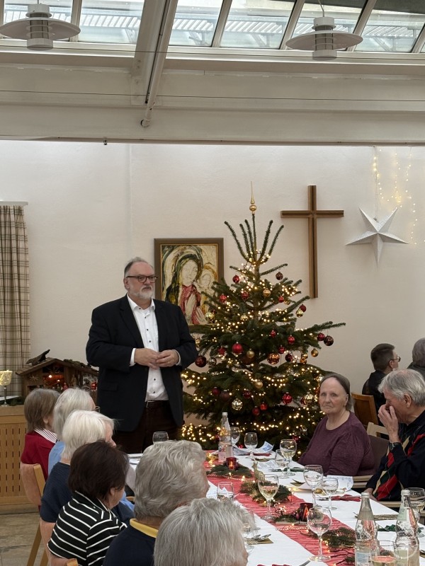 Oberbürgermeister Jürgen Dupper begrüßt die Bewohner und Gäste der Weihnachtsfeier im Seniorenstift Heiliggeist. Oberbürgermeister Jürgen Dupper steht in einem festlich geschmückten Saal und spricht zu den Bewohnerinnen und Bewohnern des Seniorenstifts Heiliggeist. Im Hintergrund ist ein beleuchteter Weihnachtsbaum sowie ein Kreuz an der Wand zu sehen. An den Tischen sitzen Seniorinnen und Senioren bei einem festlich gedeckten Essen.