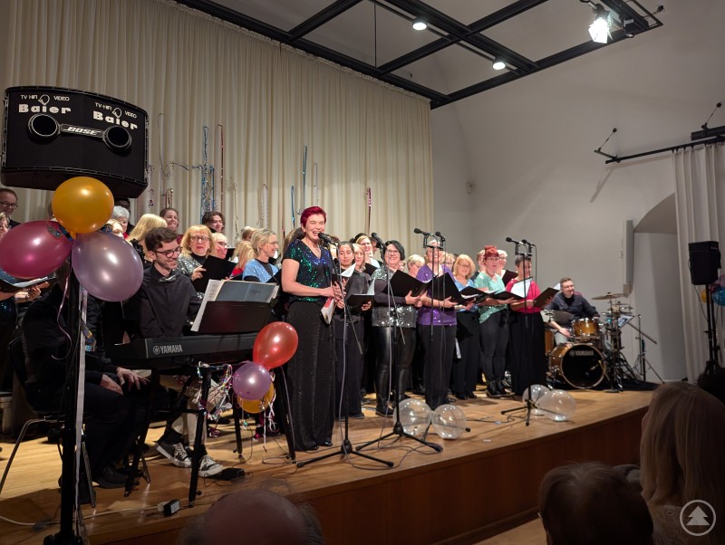 Moderatorin Beate Kern (vorne am Mikrofon) führte sympathisch und humorvoll durch den Abend. Moderatorin am Mikrofon mit Chor im Hintergrund