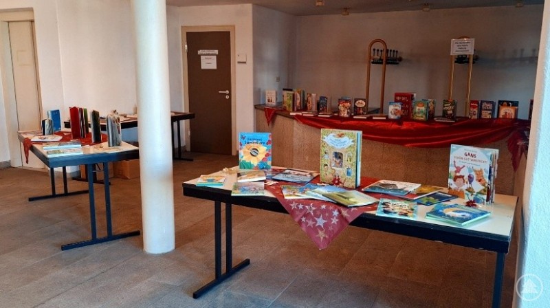Mehrere Tische sind in einem hellen Raum aufgestellt und mit einer bunten Auswahl an Kinderb&uuml;chern dekoriert. Im Hintergrund ist ein rotes Tuch &uuml;ber einen Tresen gelegt, auf dem weitere B&uuml;cher pr&auml;sentiert werden.