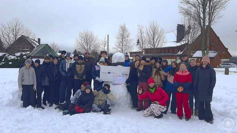 Eine gro&szlig;e Gruppe Jugendlicher und Erwachsener steht gemeinsam im verschneiten Freien, um einen Schneemann versammelt. Im Hintergrund sind schneebedeckte H&auml;user und B&auml;ume zu sehen.