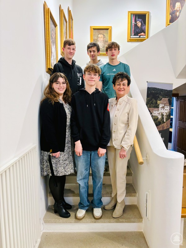 Schulleiterin Ursula Stegbauer-Hötzl re. und Museumsleitung Gabriela Rauscher li. freuen sich über die erneut gelungene Zusammenarbeit zwischen Realschule Freyung und dem Museum Jagd Land Fluss gemeinsam mit der Projektgruppe aus der Klasse 9a. Vier Schüler der Realschule Freyung stehen gemeinsam mit Schulleiterin Ursula Stegbauer-Hötzl und Museumsleiterin Gabriela Rauscher auf einer Treppe im Schloss Wolfstein und präsentieren ihr Projekt.