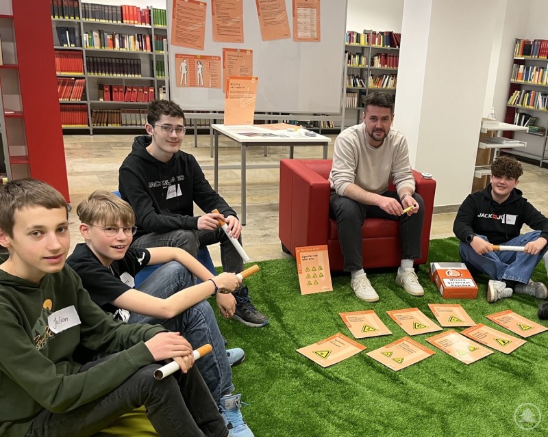 Schülerinnen und Schüler setzen sich spielerisch mit Risiken und Suchtverhalten auseinander Schülergruppe sitzt mit einem Erwachsenen auf einer grünen Fläche und beschäftigt sich mit Informationskarten