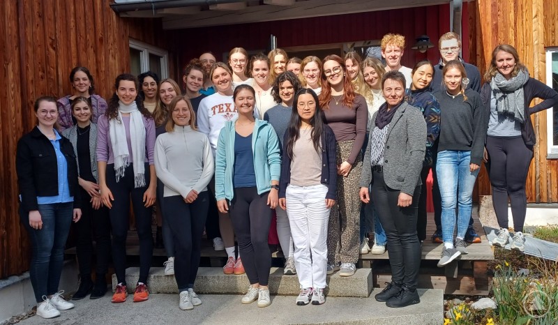 Angehende Ärztinnen und Ärzte, die derzeit in fünf Landkreisen in Niederbayern ihre Famulatur absolvieren, waren in den Kindergarten St. Josef gekommen. Das Bild zeigt eine große Gruppe junger Menschen – überwiegend Frauen – vor einem Holzgebäude. Die Stimmung ist freundlich und entspannt. Es handelt sich vermutlich um ein Gruppenfoto im Rahmen einer Fortbildung oder eines Seminars. Laut Dateiname üben hier angehende Ärztinnen und Ärzte den Umgang mit jungen Patientinnen und Patienten – möglicherweise im Rahmen eines pädagogischen oder kommunikationstrainingsbasierten Programms. Die frühlingshafte Umgebung unterstreicht die angenehme Lernatmosphäre.