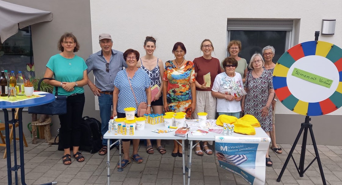 Christina Drexler (4. von links) und Alexandra Jehle (5. von rechts) gemeinsam mit Mitgliedern des Arbeitskreises Dorfladen und interessierten Besucherinnen. Eine Gruppe von Frauen und Männern steht vor einem Info-Tisch mit Sonnencreme, Flyern und Glücksrad. Im Hintergrund eine helle Hausfassade, rechts ein buntes Glücksrad mit dem Slogan „Sonnen mit Verstand“.