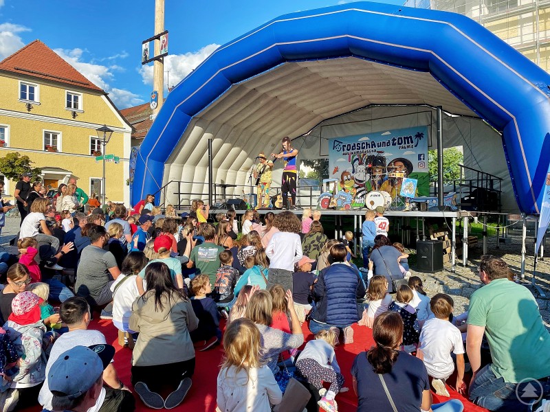 Begeisterung beim Familienkonzert in Bodenmais: Rodscha Kambodscha und Tom Palme sorgten für Musik, Bewegung und beste Stimmung. Eine große Kinder- und Familienmenge sitzt und steht vor einer Bühne mit blauer Überdachung auf dem Bodenmaiser Marktplatz. Auf der Bühne treten Rodscha aus Kambodscha und Tom Palme mit ihren Instrumenten auf und animieren das Publikum.
