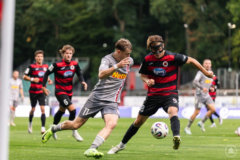 Zwei Spieler kämpfen im Zweikampf um den Ball, einer im grauen Trikot, einer im schwarz-roten Trikot.