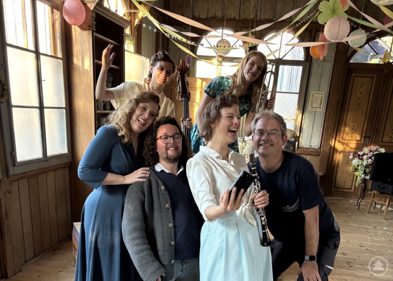 Gute Stimmung beim Dreh im Freilichtmuseum Finsterau – die „Musikgruppe 1957“ mit Regisseur Christian Lex und Komponist Florian Burgmayr. Eine Gruppe von Menschen in festlicher Kleidung posiert lachend für ein Selfie in einem dekorierten Holzsaal.