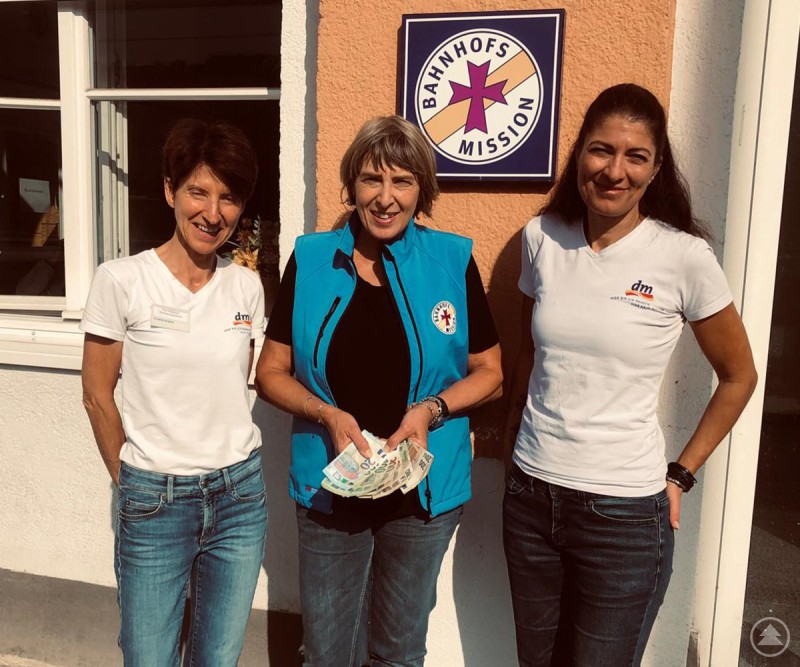 Die Marktverantwortlichen Kathrin Middeldorf (li.) und Pamela Sander (re.) übergaben die Spendensumme an Angelika Leitl-Weber, die Leiterin der Bahnhofsmission. Drei Frauen stehen vor dem Gebäude der Bahnhofsmission Passau. Zwei tragen weiße dm-T-Shirts, die Frau in der Mitte eine blaue Weste mit dem Logo der Bahnhofsmission und hält mehrere Geldscheine in den Händen.