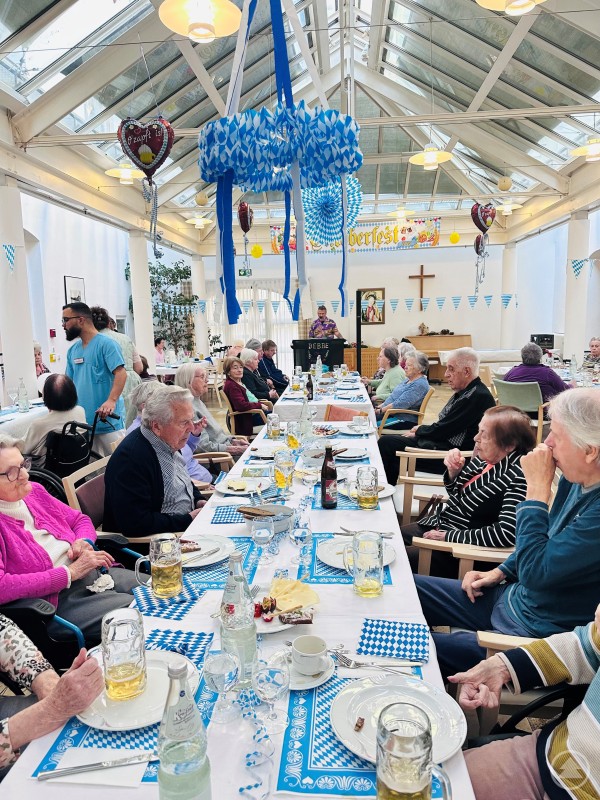 Zünftig und gesellig: Das Oktoberfest im Seniorenstift Heiliggeist bot Musik, Brotzeit und gute Laune für alle Bewohnerinnen und Bewohner. Bewohnerinnen und Bewohner des Seniorenstifts Heiliggeist sitzen an einer langen, festlich gedeckten Tafel mit weiß-blauen Dekorationen. Im Hintergrund sorgt ein Musiker für Stimmung, während Servicemitarbeiter Getränke ausschenken.