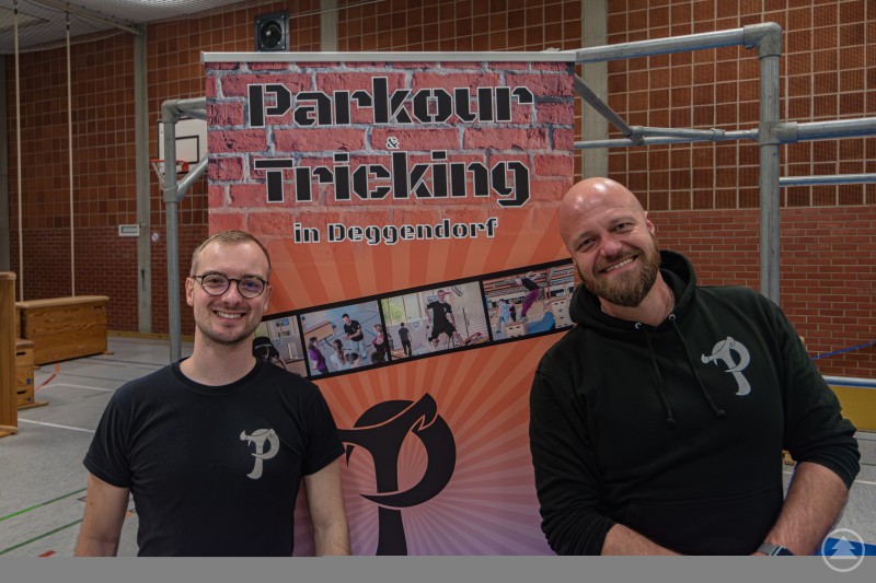 Von links Tobias Saller, neuer Leiter des Parkour-Trainings, und Ben Haslbeck vom Jugendcenter 4You. Zwei Männer stehen lächelnd in einer Turnhalle vor einem Roll-up-Banner mit der Aufschrift Parkour und Tricking in Deggendorf. Beide tragen schwarze Shirts mit einem Parkour-Logo. Im Hintergrund sind Turngeräte und eine Sporthallenwand zu sehen.