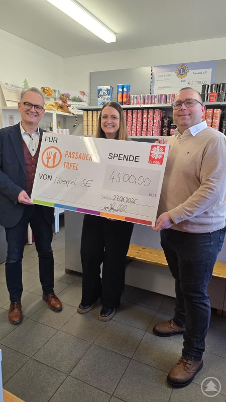Niederlassungsleiter Johannes Rahn übergibt eine Spende der Noerpel-Gruppe über 4500 Euro an Caritasvorstand Stefan Seiderer und Tafel-Koordinatorin Monika Winkler. Drei Personen stehen in den Räumen der Passauer Tafel und halten gemeinsam einen großen Spendenscheck über 4500 Euro. Hinter ihnen sind Regale mit Lebensmitteln zu sehen, die für die Ausgabe an Bedürftige vorbereitet sind.