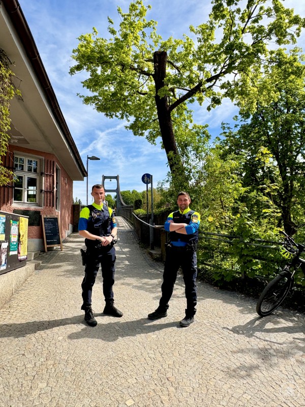 Fahrradstreife v l n r POM Leutner und PHM Winter im Einsatz in Passau. Zwei Polizeibeamte stehen auf einem Weg nahe einer Brücke und schauen in die Kamera. Sie tragen Einsatzkleidung und stehen bei sonnigem Wetter im Freien.