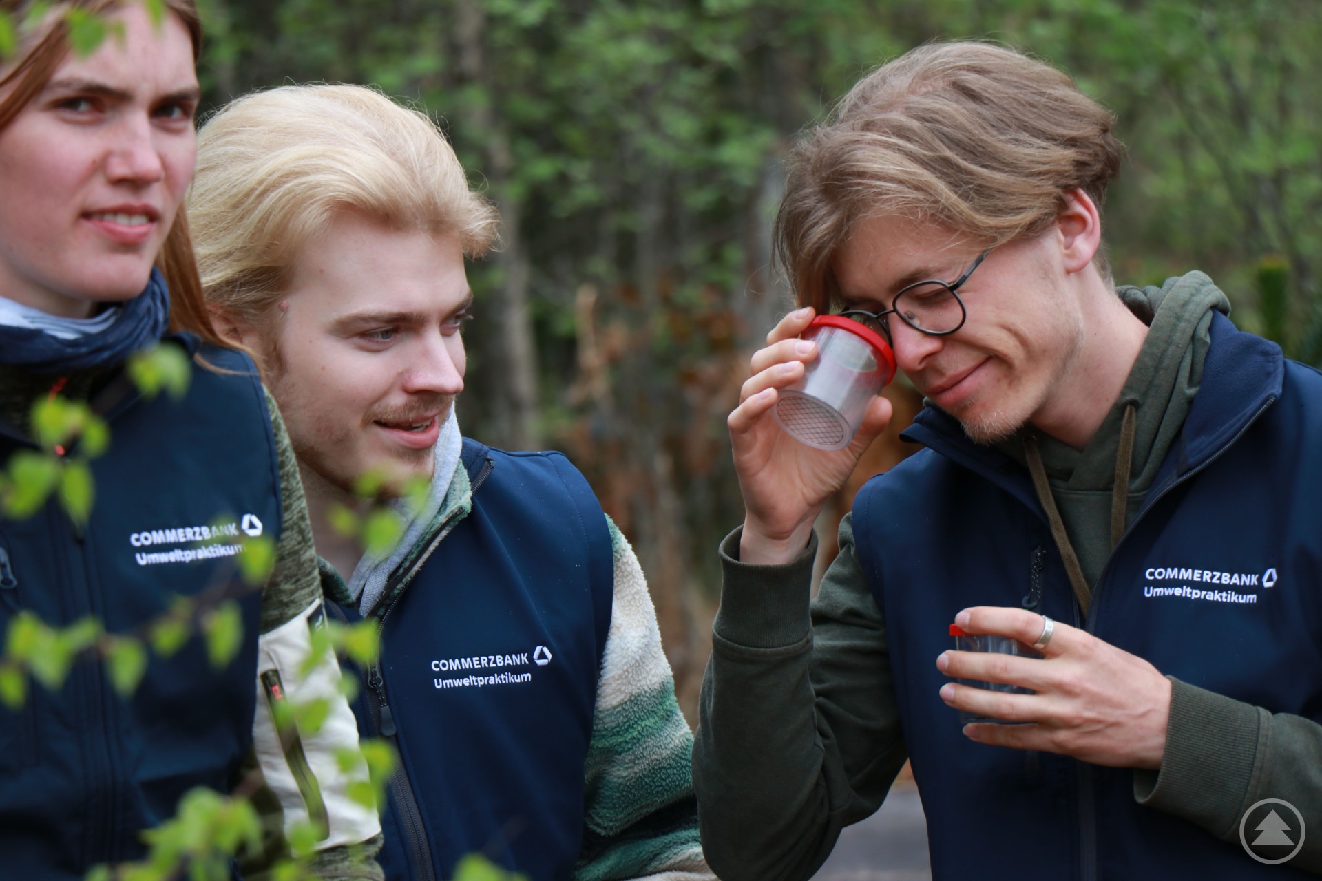 Drei junge Umweltpraktikanten im Wald, einer betrachtet ein Objekt durch eine Becherlupe.