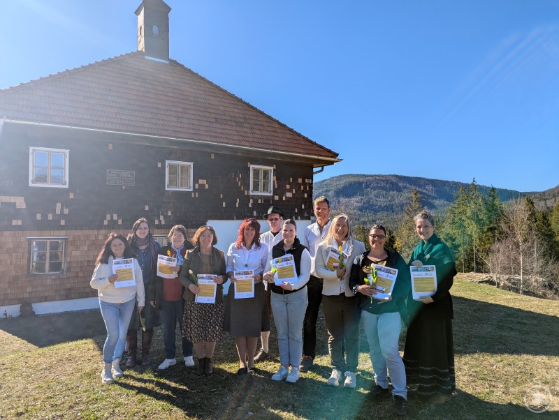 Neue Museumsbegleiter nach erfolgreich abgelegter Prüfung beim Freilichtmuseum Finsterau. Gruppe von Menschen mit Zertifikaten steht vor einem historischen Gebäude in Berglandschaft.
