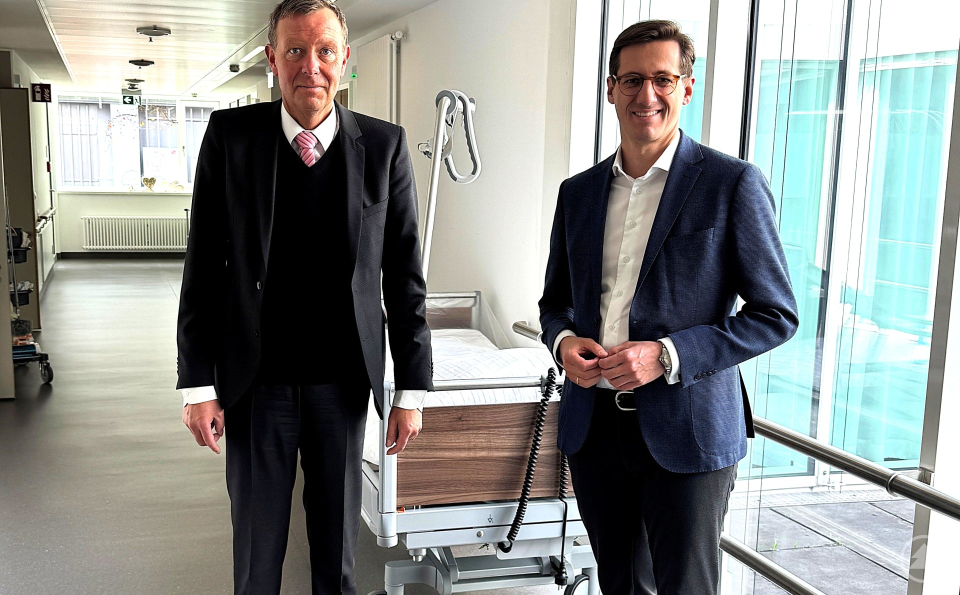 Erläuterte bei einem Rundgang durch das Haus die Erweiterungsmaßnahme am Krankenhaus Freyung: Der Geschäftsführer der Kliniken Am Goldenen Steig gGmbH, Marcus Plaschke (li.), mit dem örtlichen Stimmkreisabgeordneten Dr. Stefan Ebner (re.). Zwei Männer im Anzug stehen in einem hellen Krankenhausflur neben einem unbesetzten Klinikbett. Große Fenster lassen viel Tageslicht herein.