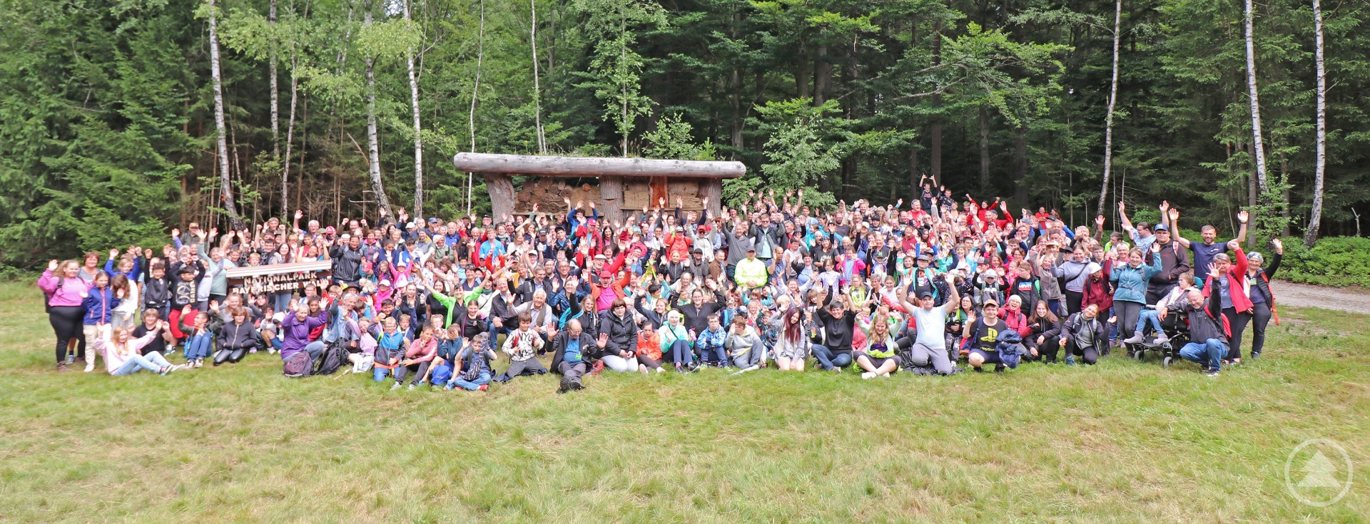 Eine große Gruppe von über 500 Menschen sitzt und steht vor einer Holzhütte im Waldspielgelände, umgeben von Wald.