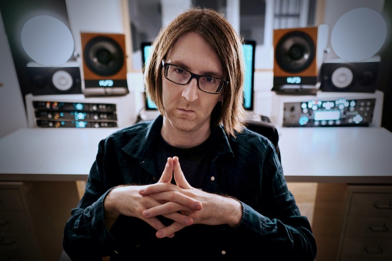 Robert Hofmann Ein Mann mit mittellangen Haaren und Brille sitzt ernst dreinblickend in einem Tonstudio vor professionellem Equipment. Er hat die Hände gefaltet und trägt ein dunkles Hemd.