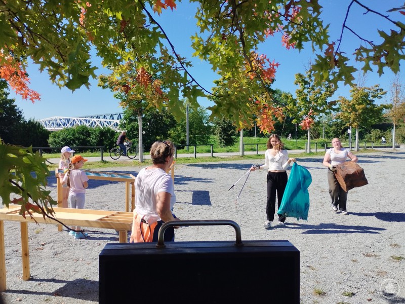 Der Weltaufräumtag am Gleisdreieck – zahlreiche Freiwillige beteiligten sich an der Sammelaktion rund um den Bogenbach. Mehrere Kinder und Erwachsene sammeln bei sonnigem Wetter Müll im Stadtpark Deggendorf. Zwei junge Frauen tragen blaue Müllsäcke und Greifzangen, während im Hintergrund ein Radfahrer vorbeifährt.