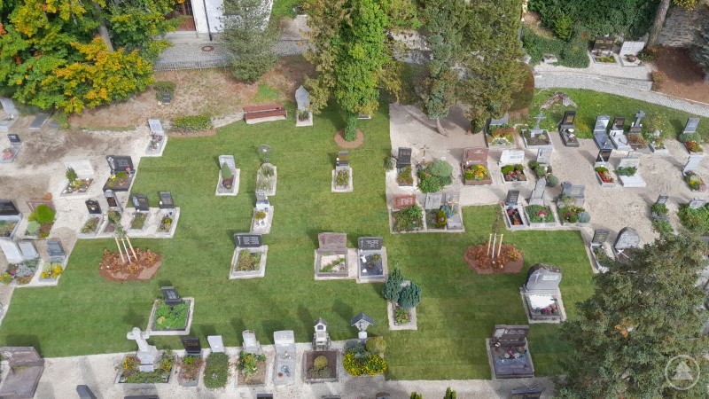 Begrünung der Friedhofskiesflächen des Ilzstadtfriedhofs. Das Foto zeigt eine Drohnenaufnahme eines Friedhofs mit gepflegten Rasenflächen, einzelnen Grabsteinen und Bäumen. In der Mitte sind neu angelegte Grünflächen mit Pflanzinseln erkennbar.