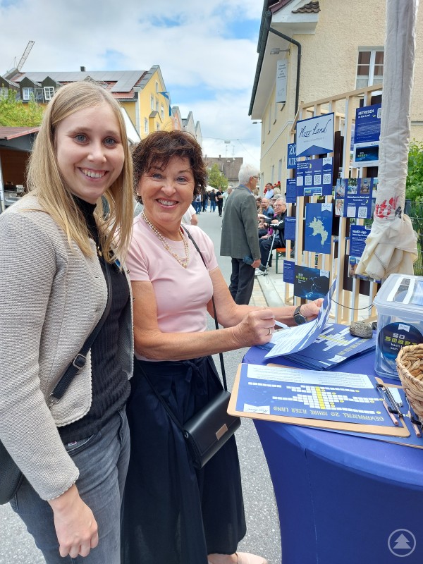 Über die Tourstopps hinweg wurde die Möglichkeit zur Teilnahme am Jubiläumsgewinnspiel rege genutzt. Im Bild Hutthurms Seniorenbeauftragte Eva Plöchinger (r.) beim Ausfüllen der Rätselkarte mit Ilzer Land Geschäftsführerin Corina Molz. Zwei Frauen stehen an einem Infostand im Freien, lächeln in die Kamera und füllen gemeinsam eine Gewinnspielkarte aus. Im Hintergrund sind Marktstände und Passanten zu sehen.