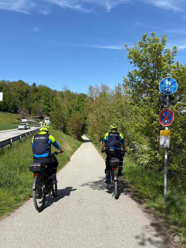 Polizeibeamte kontrollieren den Radverkehr im Rahmen der Aktion sicher mobil leben in Passau. Zwei Polizeibeamte fahren mit ihren Fahrrädern auf einem Radweg entlang einer Straße durch eine grüne Landschaft.