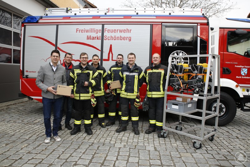 Bürgermeister Martin Pichler (v.l.), Feuerwehrbeauftragter Marktgemeinderat Walter Süß, Michael Hansbauer (Kommandant Schönberg), Tobias Loibl (Stv. Kommandant Kirchberg), Patrick Mies (Stv. Kommandant Hartmannsreit), Uli Hurzlmeier (Stv. Kommandant Schönberg) und Florian Kern (Gewählter stv. Kommandant Eberhardsreuth) mit digitalen Pagern und einem Teil der Atemschutzausstattung Das Bild zeigt Mitglieder der Freiwilligen Feuerwehr Markt Schönberg vor einem Einsatzfahrzeug. Eine Gruppe von Feuerwehrmännern in vollständiger Schutzkleidung steht gemeinsam mit einem Mann in Zivil, der eine Motorola-Schachtel hält – vermutlich bei der Übergabe neuer Ausrüstung. Rechts im Bild befindet sich ein Rollwagen mit Atemschutzgeräten und weiteren Ausrüstungsboxen.