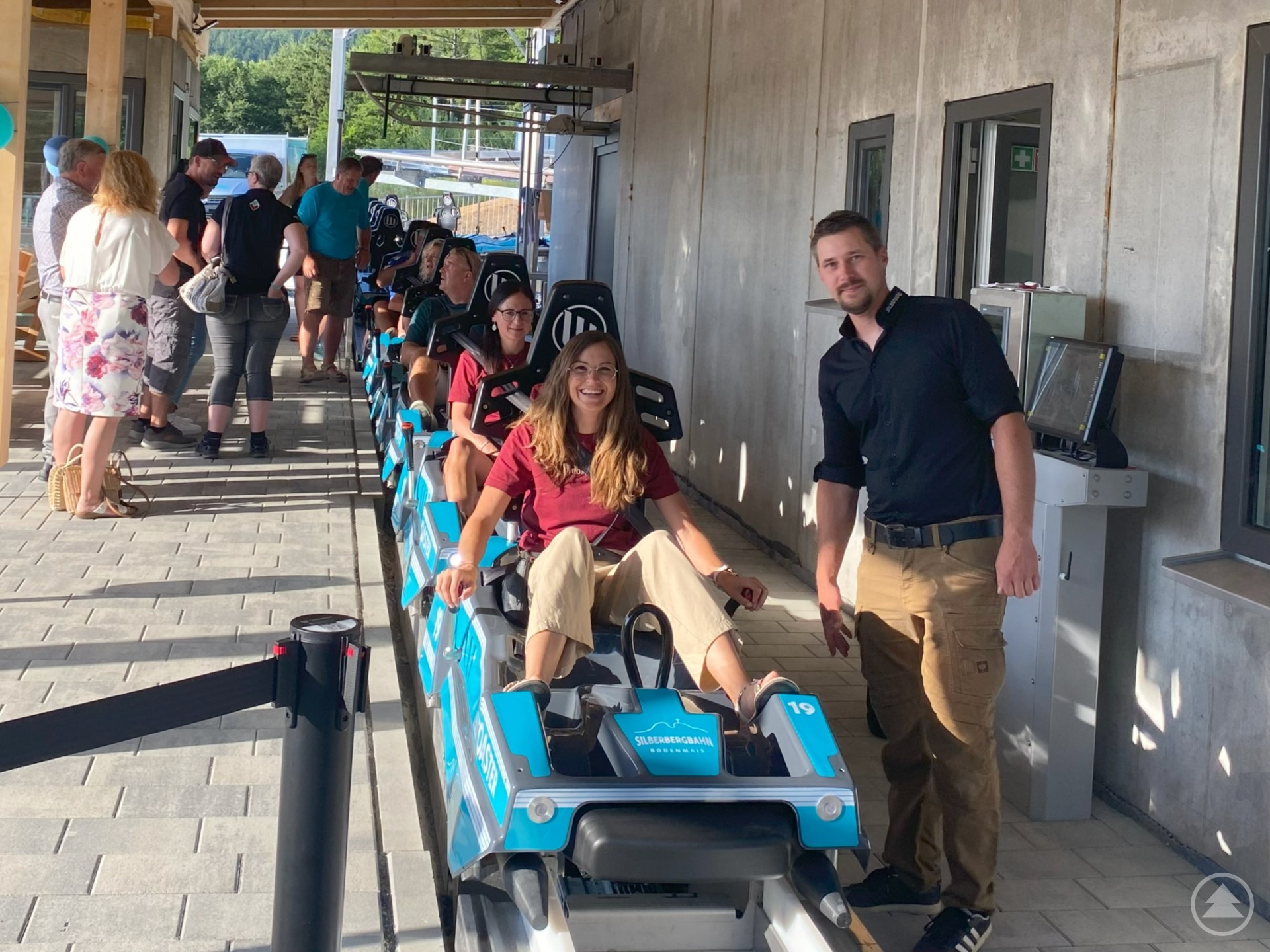 Eine Frau sitzt in einem modernen Coaster-Schlitten der neuen Allwetterrodelbahn Silberberg Coaster in Bodenmais, umgeben von weiteren Fahrgästen. Ein Mann steht lächelnd daneben. Im Hintergrund sind weitere Besucher zu sehen.