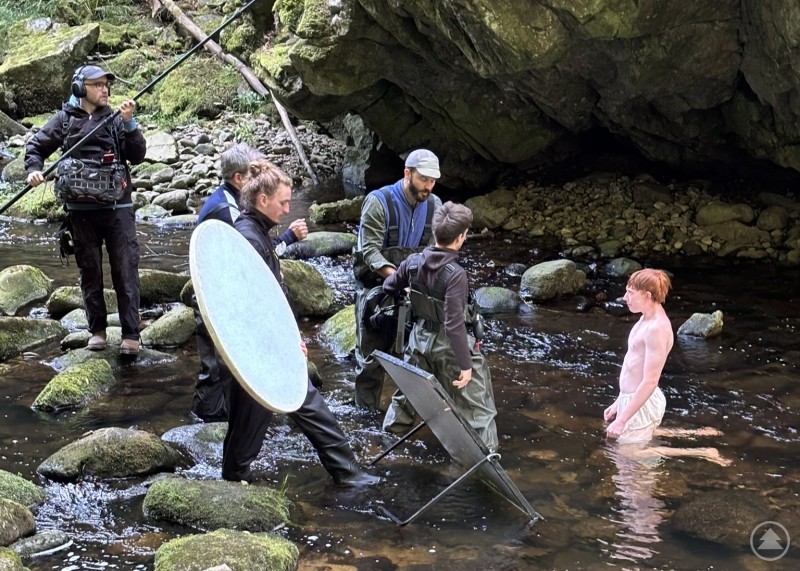 Volle Konzentration am Set – Filmcrew und Darstellerin bei Aufnahmen in der Wolfsteiner Ohe. Kameraleute und Crewmitglieder stehen mit technischem Equipment im Bach, während eine Darstellerin knietief im Wasser agiert.Leite_HYMNE (2).JPEG