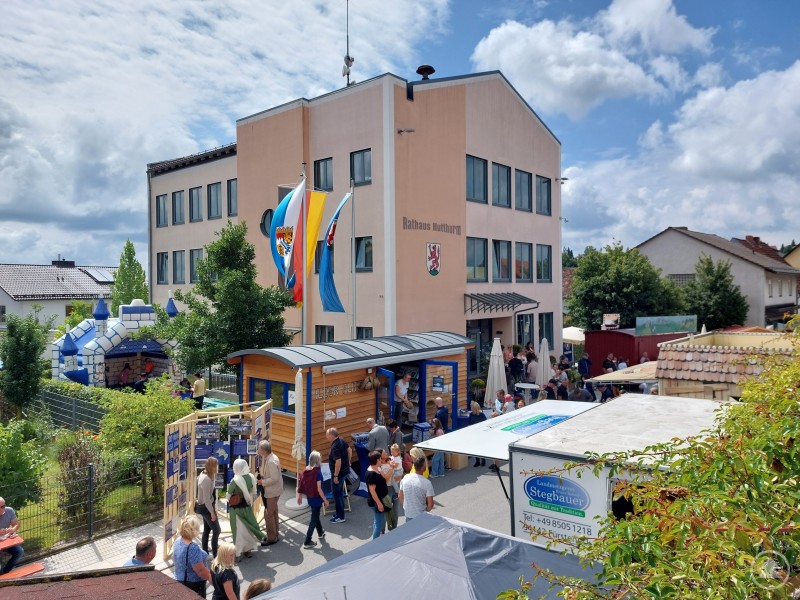 Das Ilzer Land-Team freute sich auf Tour über viel Besuch am mobilen Anhänger „Ilzer Flitzer“ und inspirierende Gespräche zur Kommunalentwicklung, wie hier beim Markttreiben in Hutthurm. Blick auf das Rathaus Hutthurm während eines Markttreibens. Viele Besucher schlendern zwischen Ständen und Informationszelten, im Hintergrund wehen Fahnen und eine Hüpfburg steht im Gartenbereich.