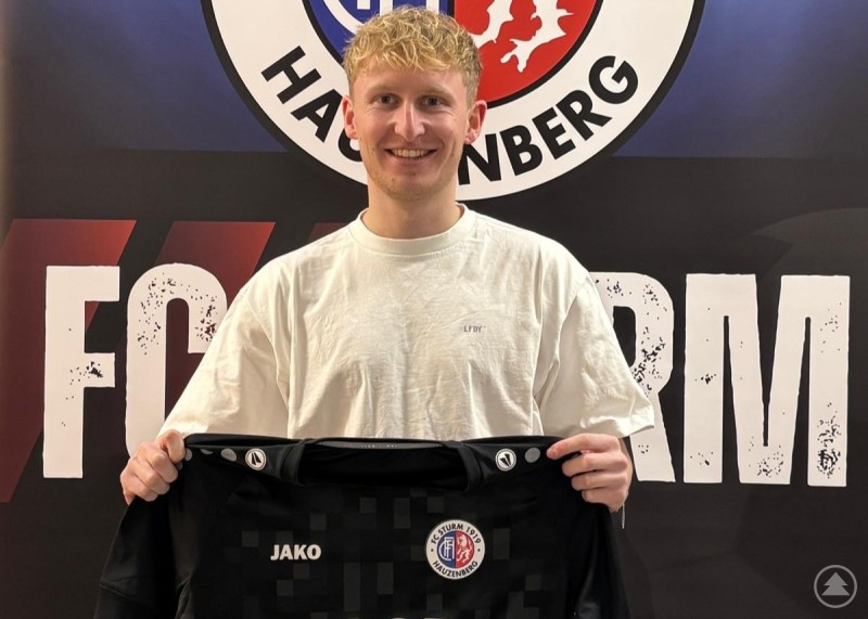 Jonas Windpassinger verstärkt den FC Sturm Hauzenberg zur kommenden Saison. Ein Fußballspieler präsentiert lächelnd ein neues Trikot des FC Sturm Hauzenberg vor dem Vereinslogo.