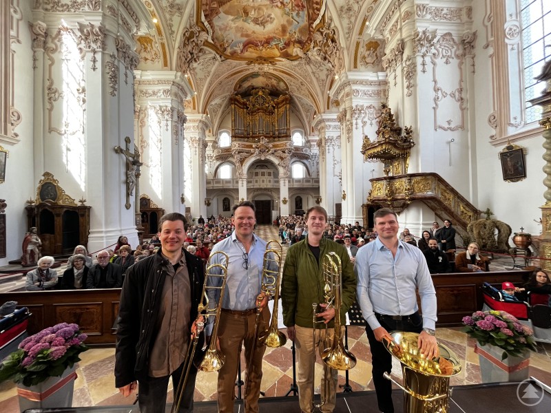 Die European Orchestra Academy begeisterte auch bei öffentlichen Konzerten, unter anderem in beeindruckender Kulisse vor großem Publikum. Vier Musiker mit Blechblasinstrumenten stehen in einer prunkvollen Barockkirche vor einem großen Publikum und lächeln in die Kamera.