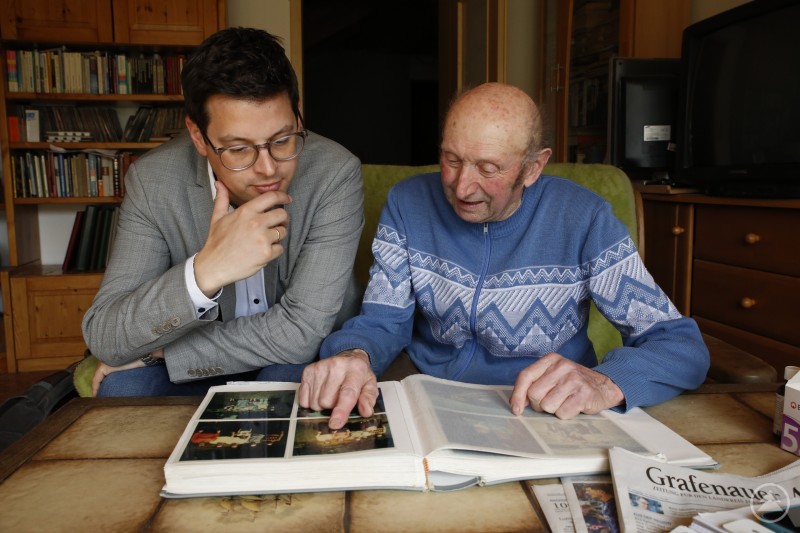 Bürgermeister Martin Pichler war überrascht, wie viele Erinnerungen sich Josef Peter bei seinen Märschen durch die Region eingesammelt hat. Ein Schatz, der viel Vergangenheit unvergesslich macht. Ein älterer Mann und ein jüngerer Mann blättern gemeinsam in einem alten Fotoalbum in einem Wohnzimmer voller Bücher und Holzschränke.