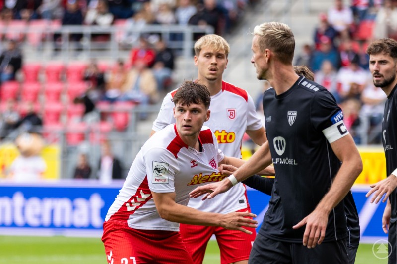 ine Spielsituation im Stadion. Spieler in weißen Trikots mit roter Hose (mit dem Schriftzug „Netto“ als Sponsor) stehen einem gegnerischen Team in schwarzen Trikots gegenüber. Ein Spieler im Vordergrund wirkt fokussiert und bereit, sich durchzusetzen.