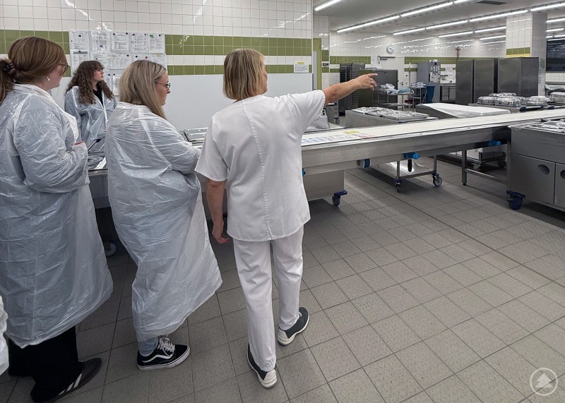 Mehrere Personen in Schutzkleidung stehen in einer Großküche, wo eine Mitarbeiterin auf verschiedene Arbeitsbereiche zeigt.
