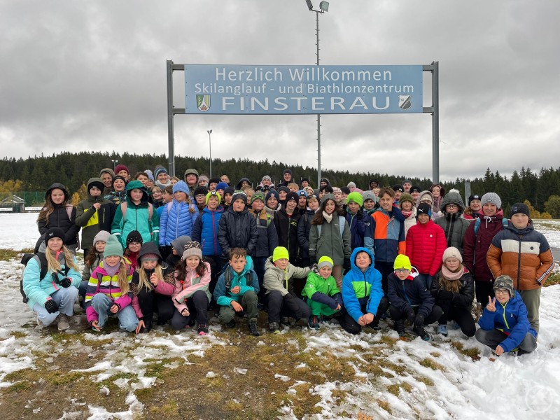 Trotz des überraschenden Wintereinbruchs hatten die Fünftklässler der Realschule Grafenau im Schullandheim in Finsterau viel Spaß. Eine große Gruppe von Schülerinnen und Schülern steht mit ihren Lehrkräften vor einem Schild des Skilanglauf- und Biathlonzentrums Finsterau. Im Hintergrund liegt Schnee, und die Kinder tragen bunte Winterjacken und Mützen.